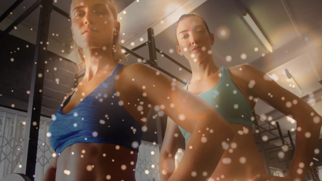 Two women posing, camera tilting up revealing faces for fitness marketing with foreground particles