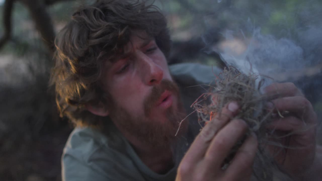 수염이 있는 백인 남성 생존주의자 는 야생 에서 캠프 에 불 을  ⁇ 기 위해 담배 를 피우는 연료 를 불어 넣고 있다