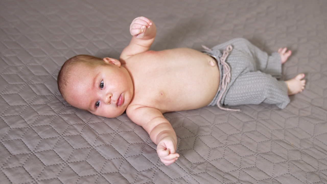 Adorable little boy wearing grey pants only lies on the bed. Plump small sweet baby in pants on the grey backdrop.