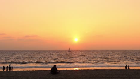 Hermoso-Atardecer-En-La-Playa-Con-Un-Bote-Navegando-En-El-Océano-Y-Gente-Caminando-En-La-Arena-Mirando-Las-Olas