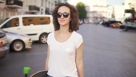 happy woman touching her sunglasses then starting to walk holding her stylish longboard in the morning. slow motion shot