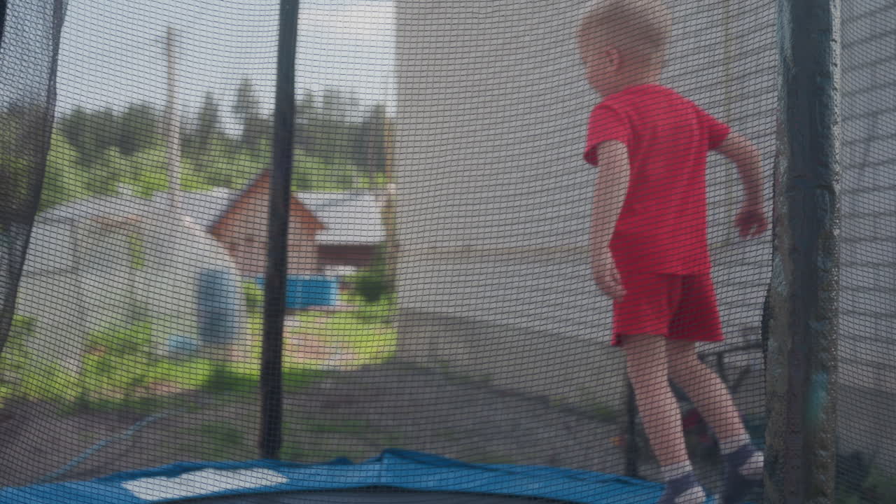 Little Toddler In Vibrant Red Romper Enjoys Lively Trampoline Fun Outdoors, An Energetic Young Child In Cheerful Red Romper Enthusiastically Bounces On Backyard Trampoline During Summer