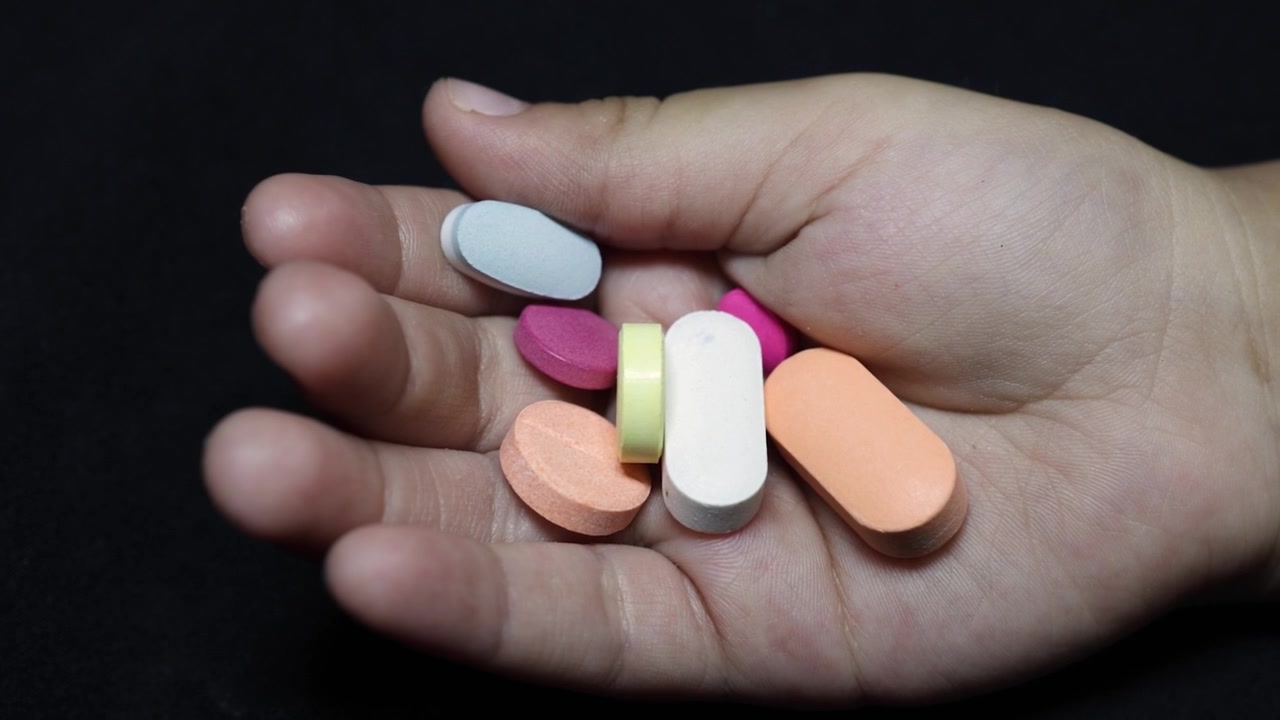 Palma da mão de criança cheia de comprimidos coloridos de medicamento em fundo escuro.