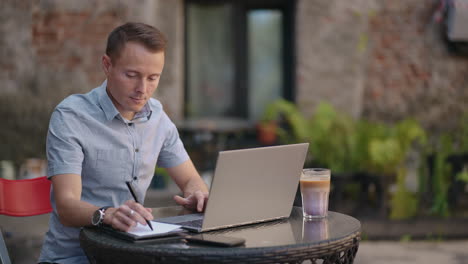 Ein-Mann-Lernt-Aus-Der-Ferne,-Auf-Einen-Laptop-Zu-Schauen-Und-Schreibt-Mit-Einem-Stift-In-Ein-Notizbuch-Oder-Notizbuch