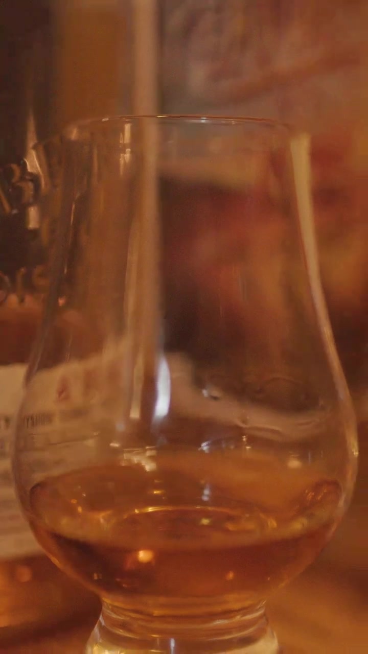 Una scena calda e delicatamente illuminata con una lenta panoramica della telecamera su bicchieri da degustazione di whisky su un tavolo di legno, mettendo in risalto il liquido ambrato e un'atmosfera accogliente.