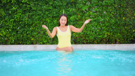 smiling young asian woman is playing with water while sitting on pool border on hot summer day, slow motion