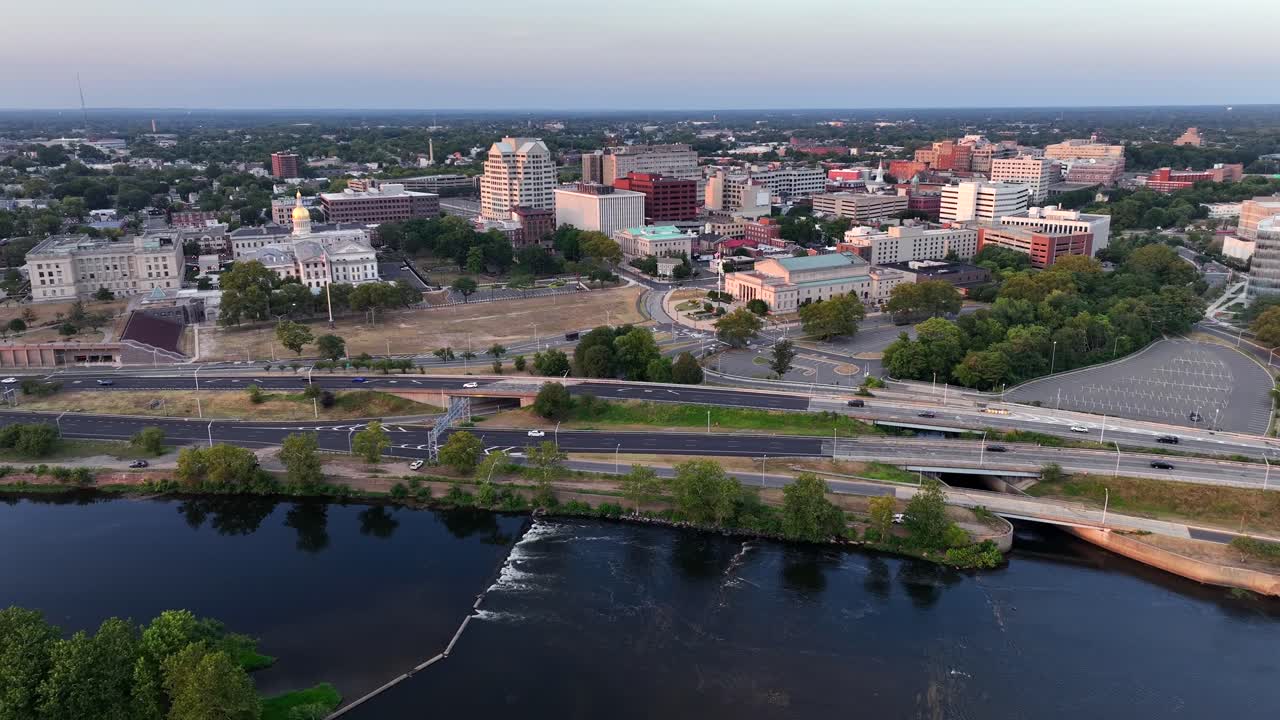 Traffic on coastal road in front of Trenton Downtown at sunrise. Descend drone wide shot. Summer day in New Jersey. High-rise buildings and offices in district. Financial area