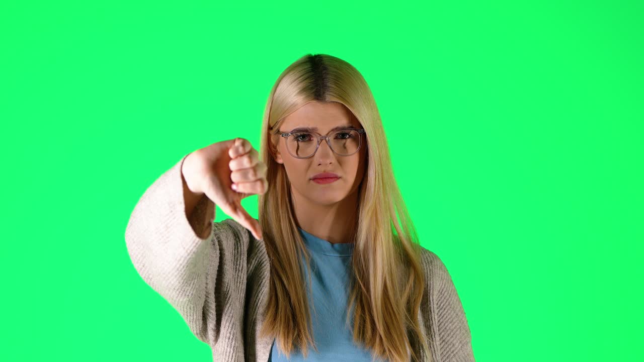 Smart looking blonde caucasian woman thumbs down with negative attitude looking sad at camera, portrait shot in green chroma infinite background studio
