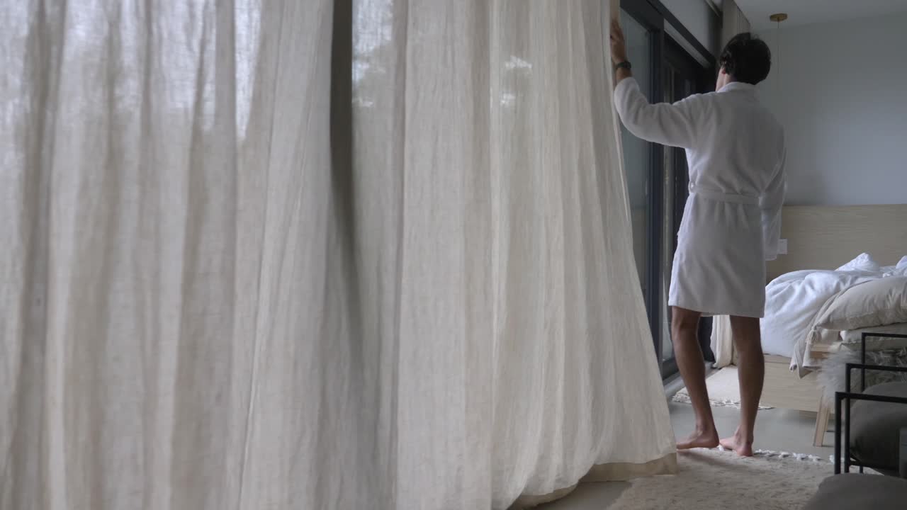 A Man Wearing Bathrobe Is Closing Curtain Of A Glass Window Wall