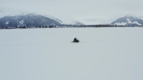 man on snowmobile traveling on snowy field with mountains in background, aerial