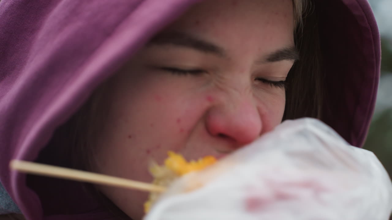 겨울 재을 입은 젊은 여자가 거리에서 옥수수를 즐기고, 먹는 동안 미소 짓고, 후드 재, 추운 날씨, 야외, 눈과 나무로 흐릿한 배경