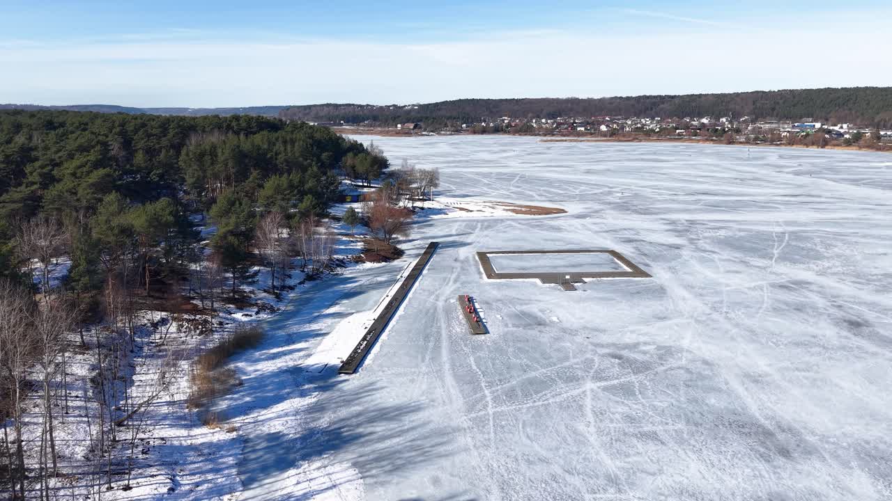 Záběry z dronu ukazující rozlehlé ledové jezero, ohraničené hustým, zasněženým lesem pod jasně modrou oblohou. Ideální pro zimní, přírodní a sezónní témata, zachycené v ohromujících detailech.