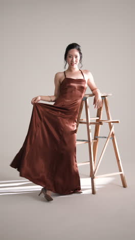 vertical video: elegant woman in satin dress posing thoughtfully against wooden ladder in studio