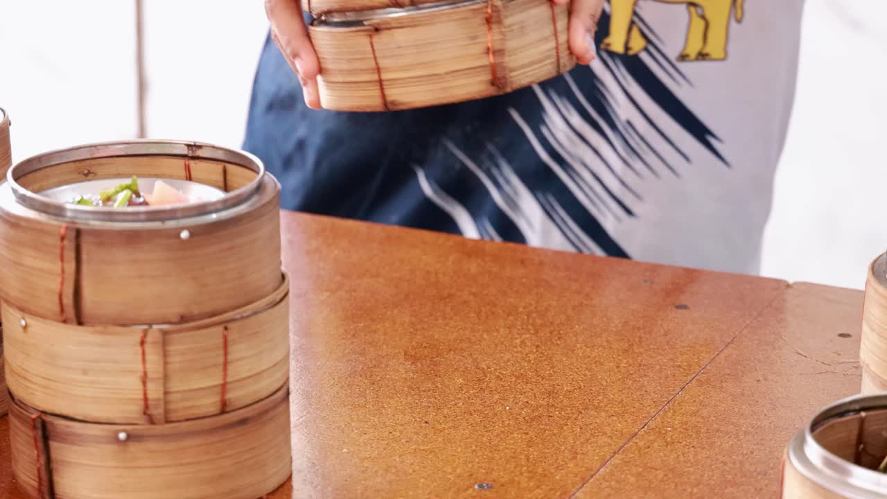 Hands arrange bamboo steamers filled with dim sum on a wooden table in bright lighting