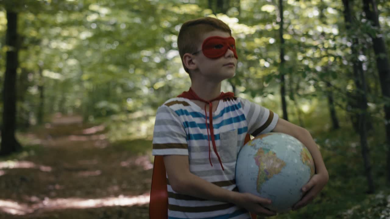 chico caucásico rojo con traje de superhéroe sosteniendo un globo y caminando por el bosque.