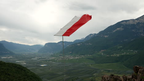 Fuerte-Viento-Ondeando-La-Bandera-Roja-Y-Blanca-Del-Castillo-Leuchtenburg-Kaltern-Durante-El-Día-Nublado-En-Italia-Entre-Montañas-Y-Valle