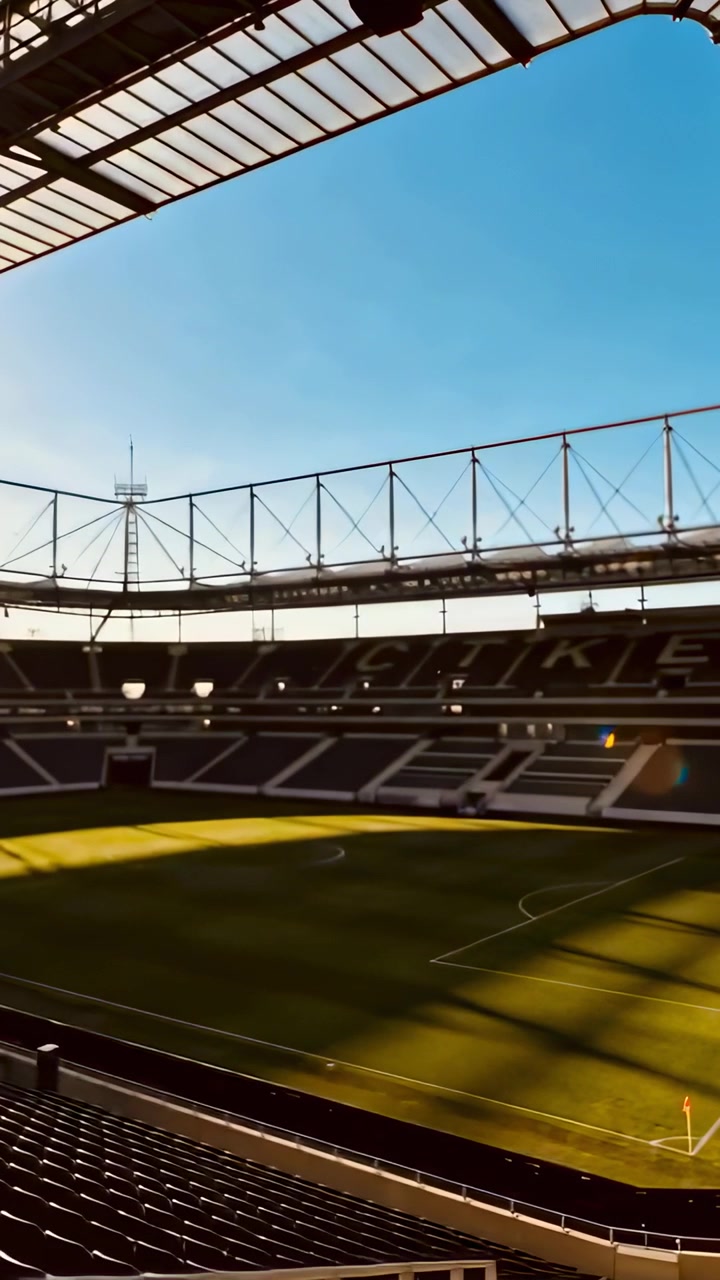 Vertikales Video: Neigender Grashalm enthüllt leere Stadiontribüne mit sonnenbeschienenem Spielfeld, Lens Flare