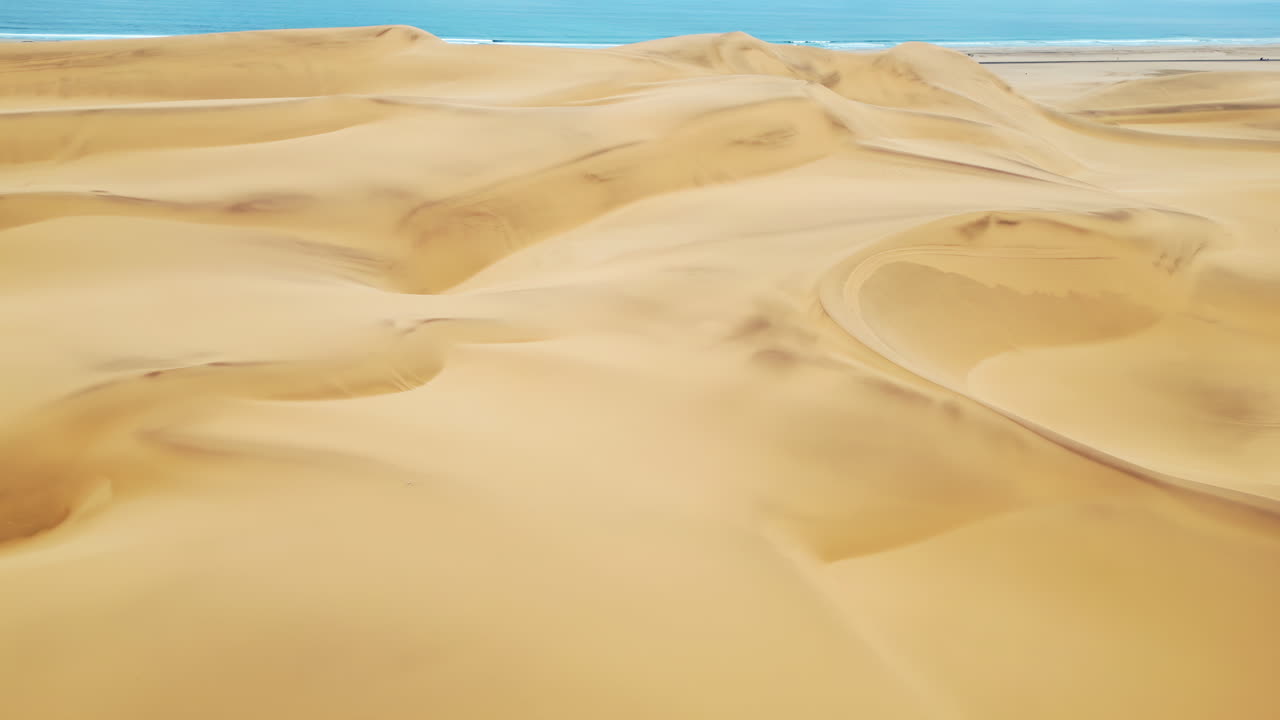 Vista aerea con drone delle dune di sabbia dorata del Deserto del Namib con tracce di pneumatici che attraversano le dune e l'Oceano Atlantico visibile in lontananza