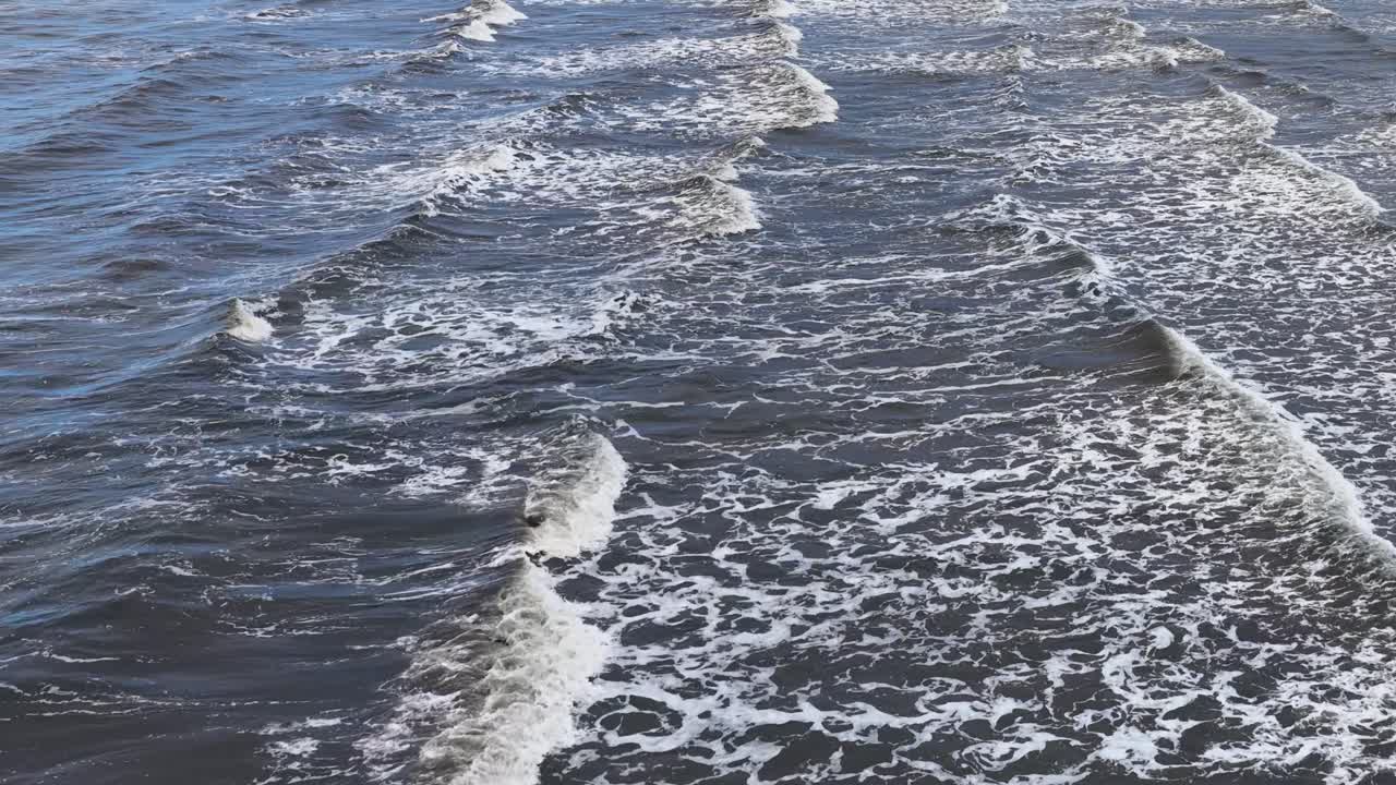 Shallow, muddy waves break in parallel lines over a murky shoreline, viewed from above in soft daylight with steady, static camera framing