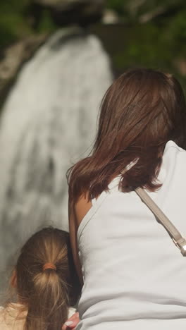 mother and little daughter enjoy waterfall view in wild nature park. loving girl leans on woman looking at water flow cascade at reserved area