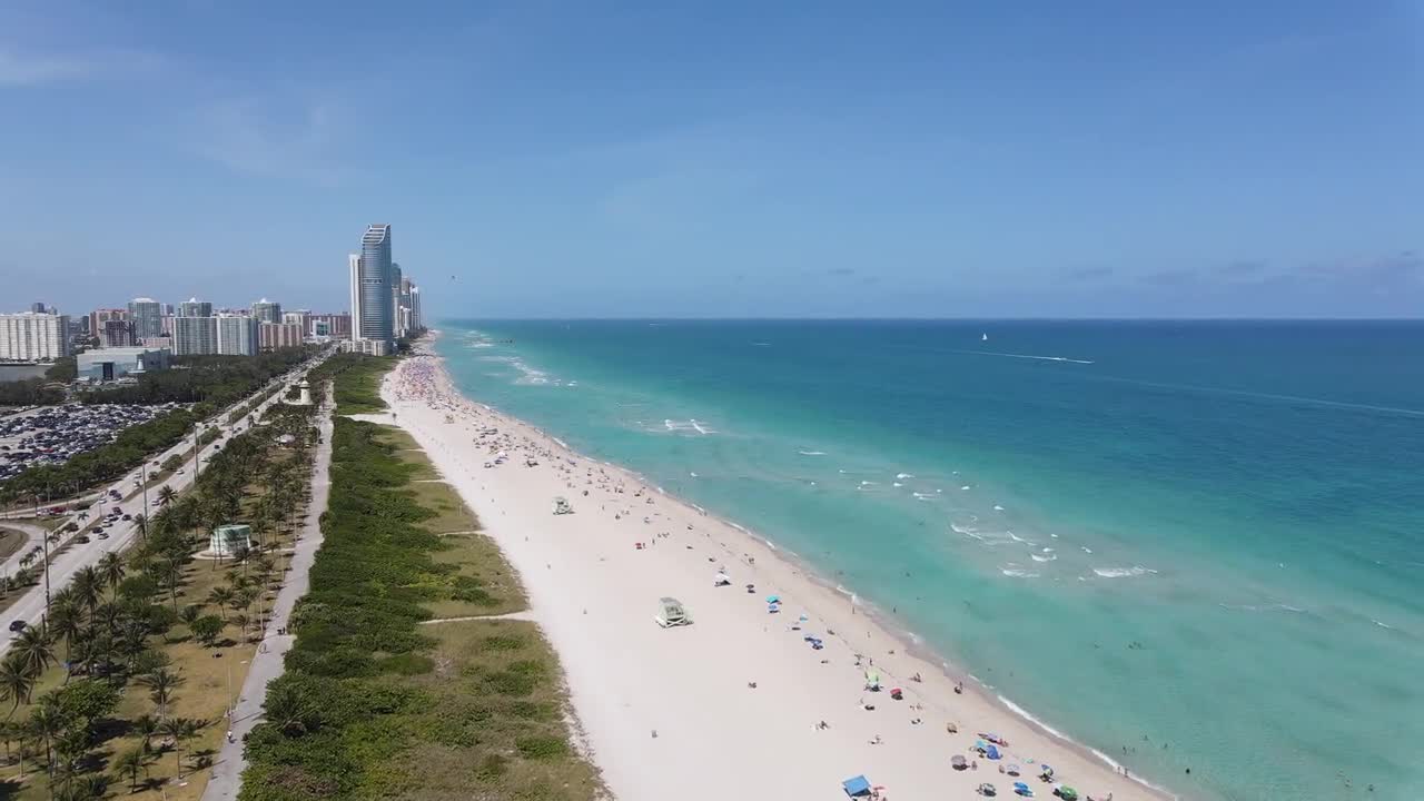 A stunning aerial view of a wide, sandy beach lined with turquoise waters, dotted with beachgoers and bordered by a scenic road with palm trees. High-rise buildings overlook this sunny paradise.