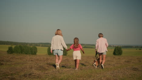 mutter und tochter gehen hand in hand, während der haustierliebhaber das seil hält, und ihr aufgeregter hund geht eng neben ihr auf einem weit offenen feld, umgeben von fernen bäumen und üppigem grün, unter klarem himmel