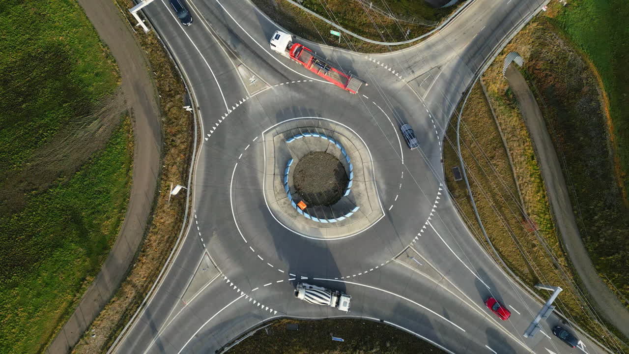 Drone shot looking straight down at a multi-lane roundabout with cars and trucks circulating smoothly, surrounded by rural roads and fields