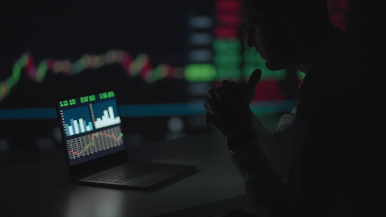 Late at Night in Private Office Businessman Works on a Laptop. Late at Night Trader Reads Numbers on His Multiple Displays with Stock Information on Them