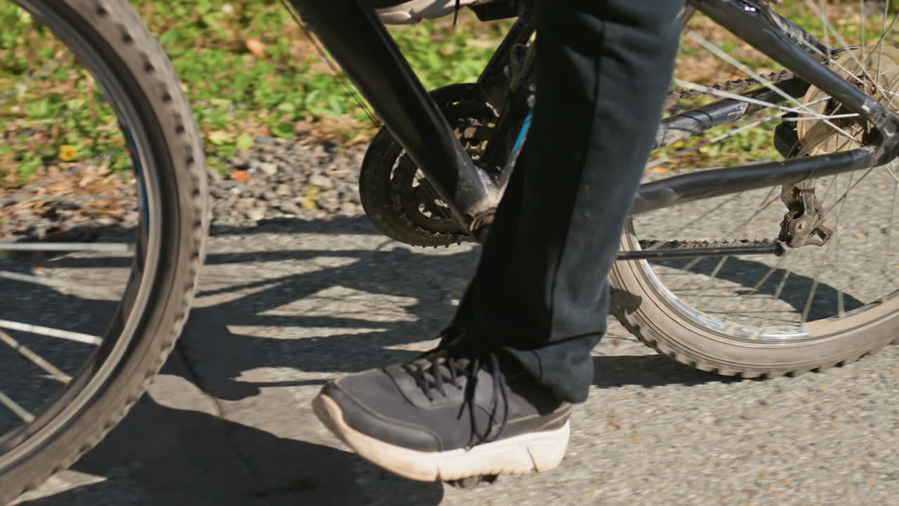 Close de uma roda de bicicleta e um pé pedalando; entregador de bicicleta, de jeans e tênis casuais, andando no asfalto iluminado pelo sol; corrente e pneu com poeira, sombra projetada no pavimento, transmitindo movimento constante e último urbano