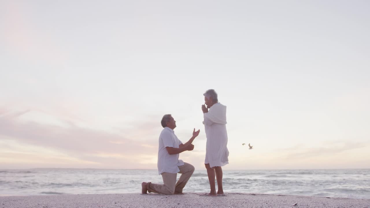 히스패닉 성인 남자가 해가 지는 해변에서 성인 여성에게 청혼하고 있습니다.