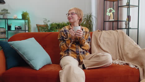 Attractive smiling young woman drinking a cup of coffee or herbal tea relaxing resting at home couch