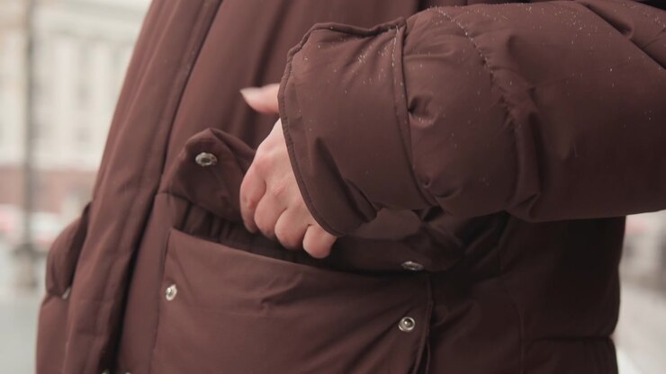 Torso and arm in dark brown padded coat with hand sliding into pocket, wet droplets and damp patches on sleeve and pocket, cool urban outdoor light, winter commute mood, with coat, pocket, hand