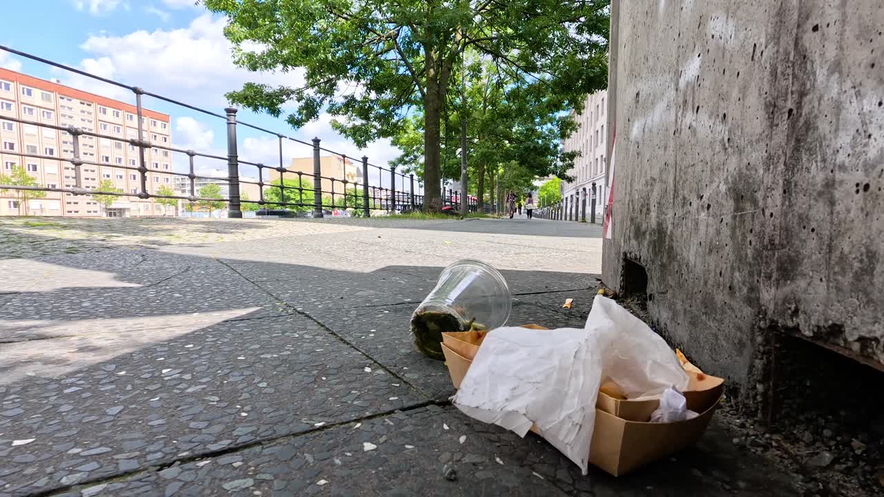 A person walks past discarded food packaging and trash on a city sidewalk in Berlin, with bright daylight and static low-angle camera framing