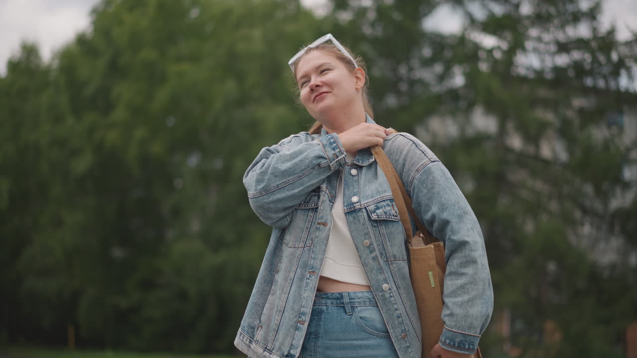 Hvid kvinde i denimjakke smiler, spadserer gennem løvfyldt park mens hun justerer vævet mulepose, kigger op på overskyet himmel med afslappet selvsikkert udtryk, ægte portræt i naturligt lys, roller.