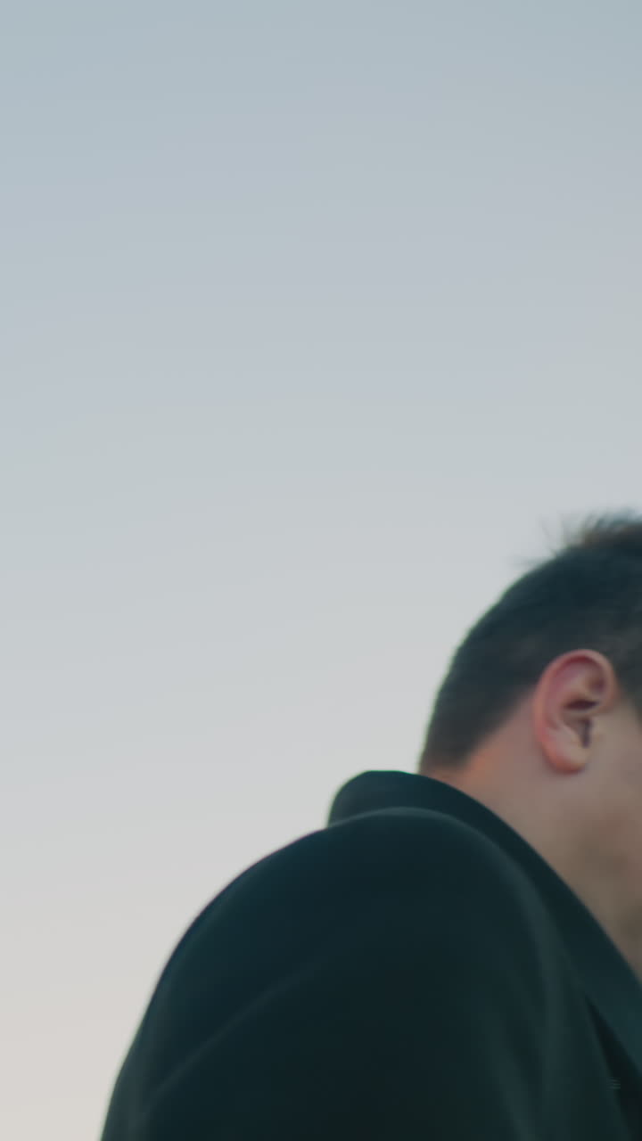 검은 코트와  셔츠를 입은 자신감 넘치는 남자가 부드러운 하늘 아래 밖에서 드폰에 초점을 맞추고, 휴대전화를 주머니에 돌려놓는 미묘한 움직임은 도시 기업 환경에서 평온한 결단력을 반영합니다.