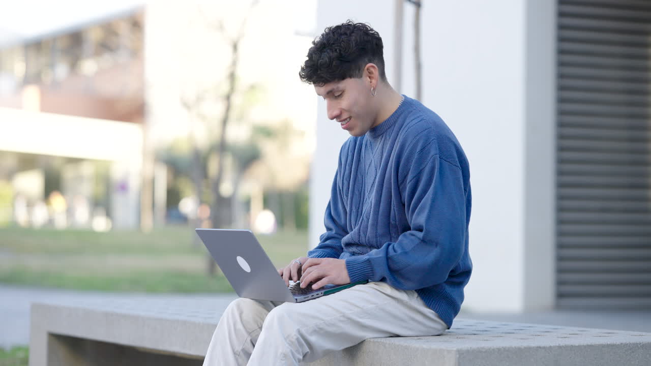 Young hispanic student working on his laptop sitting on a bench