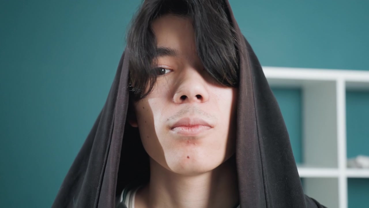 Pria Asia di rumah dengan handuk pembersih wajah. Pria muda Asia terbungkus handuk berkerudung di rumah, mengaplikasikan pembersih wajah dan memeriksa kulit dengan latar belakang rak berwarna teal. Momen kamar mandi yang tenang dan intim menunjukkan