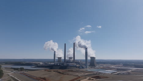 static aerial drone video of a large power plant with multiple smokestacks and cooling towers releasing steam into a clear blue sky in georgia.