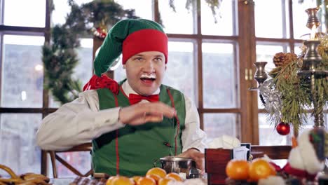 Funny elf with drinks and becomes dirty on his face on Christmas background. Hungry male elf in red and green costume drinks at festive table indoors. Close-up.