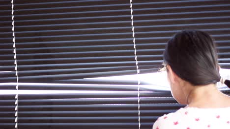 Young-woman-looking-through-window-with-blinds-and-bright-light-coming-through