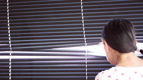 Young woman looking through window with blinds and bright light coming through