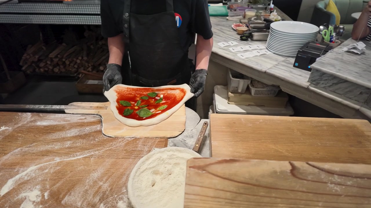 Siyah önlüklü şef, ticari bir mutfakta ahşap bir kürek üzerinde taze Margherita pizza hazırlıyor.
