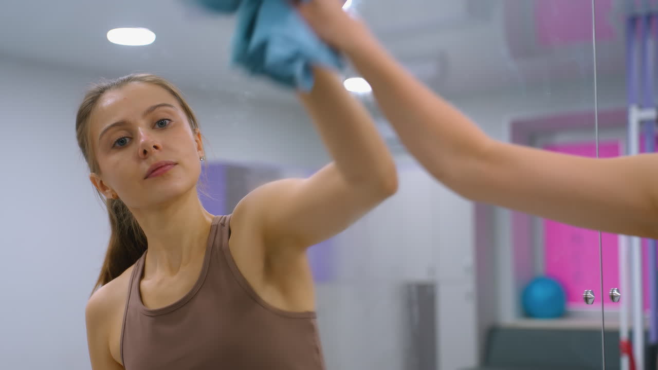 갈색 스포츠 옷을 입은 여성은 현대 체육관의 큰 거울에 청소 용액을 스프레이합니다. 거울은 체육관 장비, 분홍색 창문 개, 밝은 조명을 반영합니다.