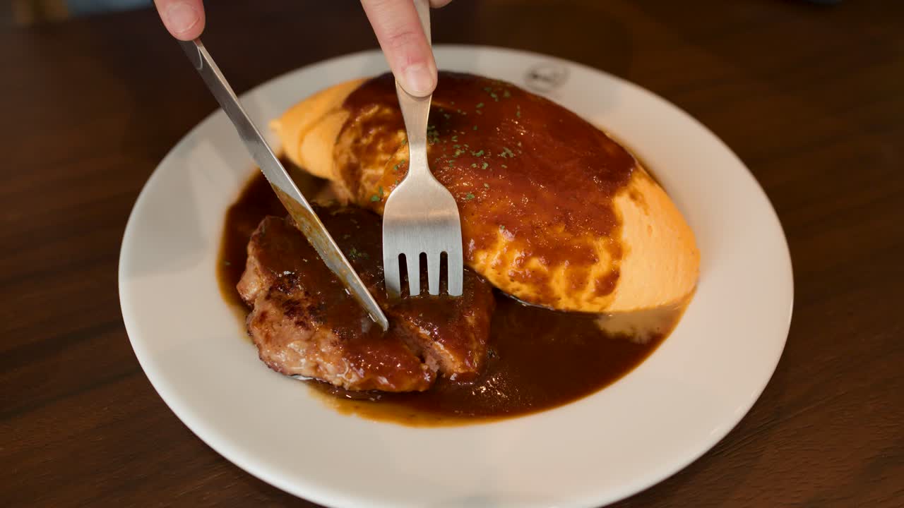 Hand slices hamburg steak beside omelette rice, brown sauce, overhead view, warm indoor lighting