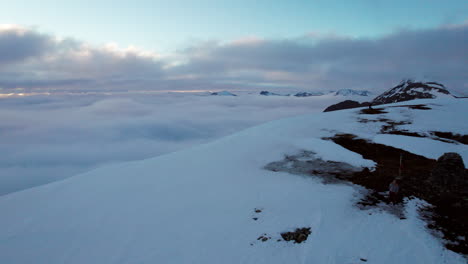 Sobrevuelo-Aéreo-De-Un-Montañero-Parado-En-El-Borde-De-Un-Acantilado-Cubierto-De-Nieve-Que-Revela-Un-Paisaje-Cubierto-De-Nubes,-Con-Un-Sol-Bajo