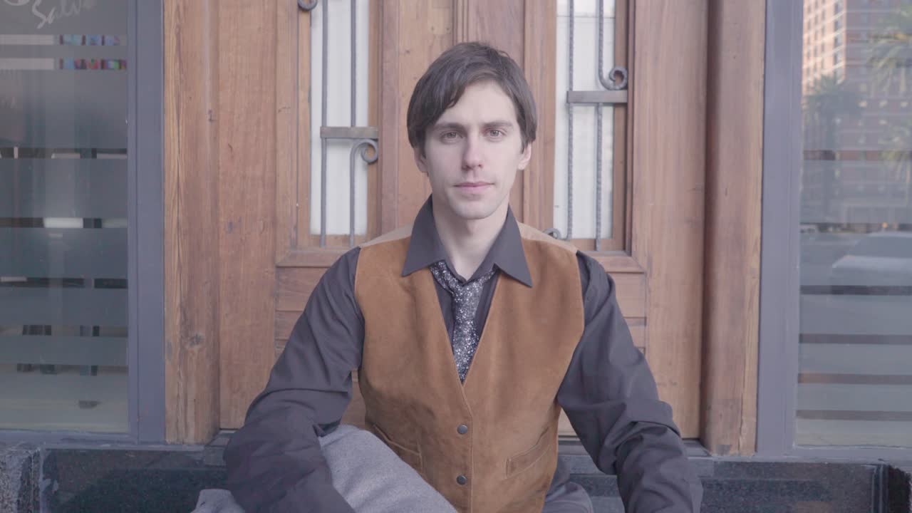 Young man in shirt and tie sitting on the sidewalk, looks at the time on his wrist watch and then looks at camera