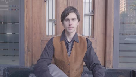 young man in shirt and tie sitting on the sidewalk, looks at the time on his wrist watch and then looks at camera