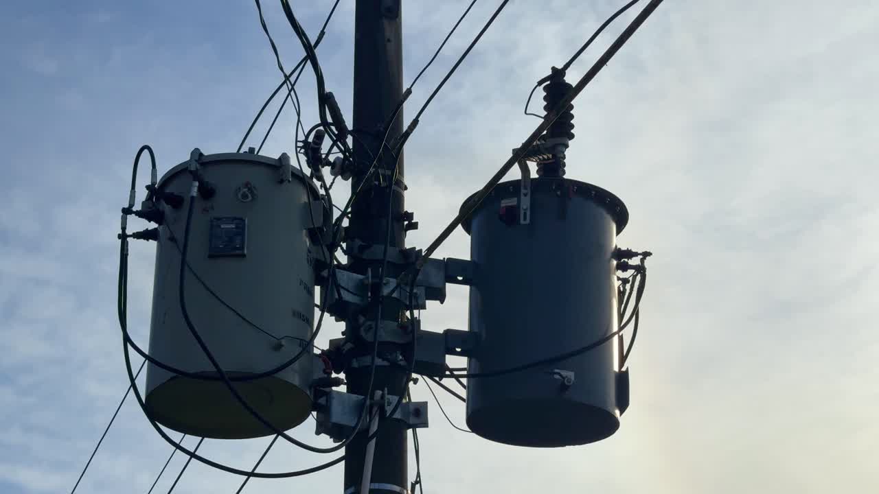 Utility pole-mounted transformer system with insulated wires and metal fittings, shown against a clear daylight sky, representing energy infrastructure