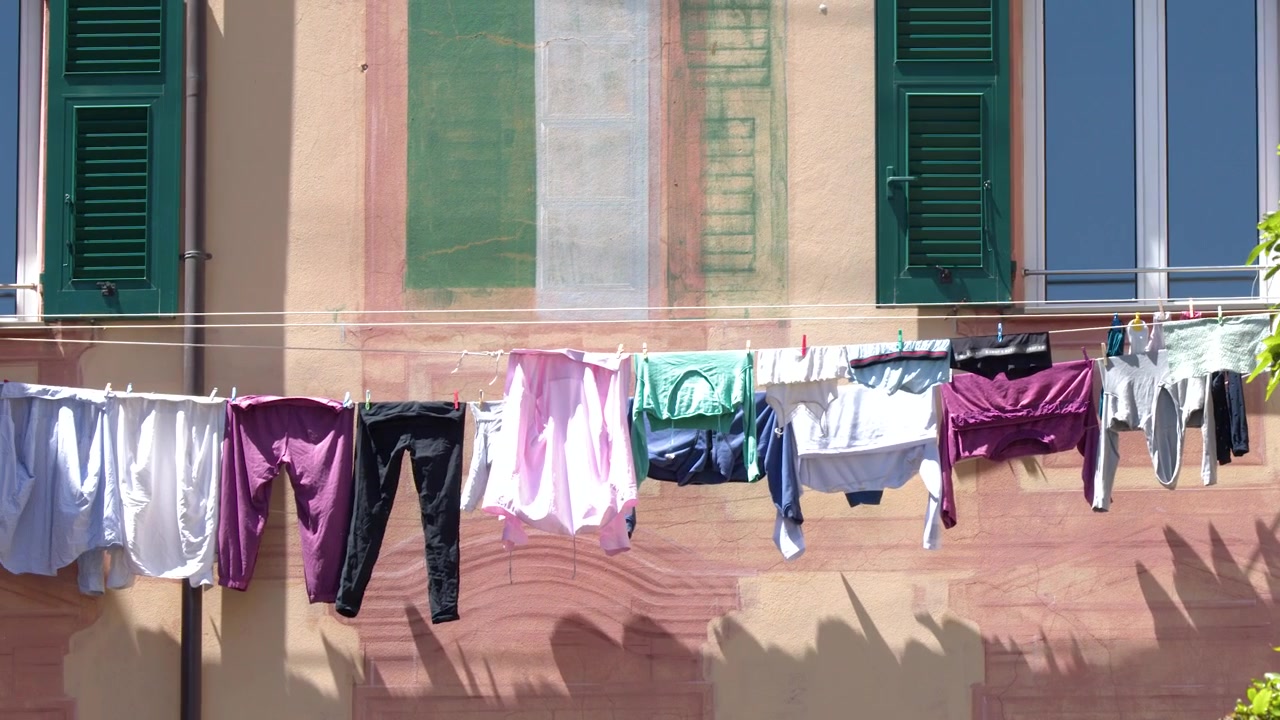 Colorful clothes dry on a line against a pink building with green shutters in Italy.