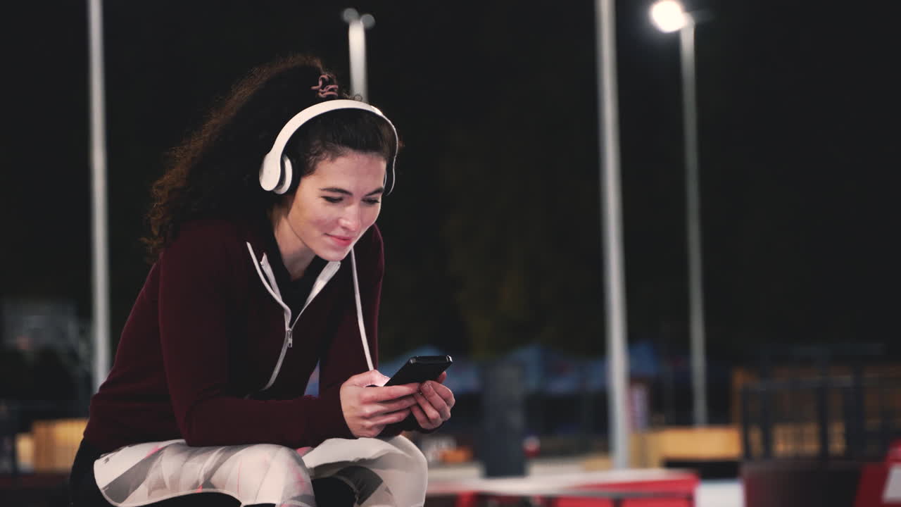 mujer deportiva sonriente sentada en el parque escuchando música con auriculares bluetooth y enviando mensajes de texto en su teléfono móvil mientras toma un descanso durante su sesión de entrenamiento por la noche